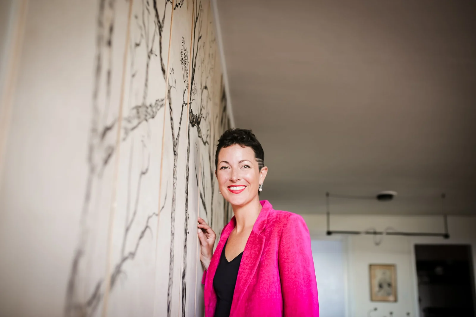 Louise laughing in a bright pink suit beside her wooden forest mural